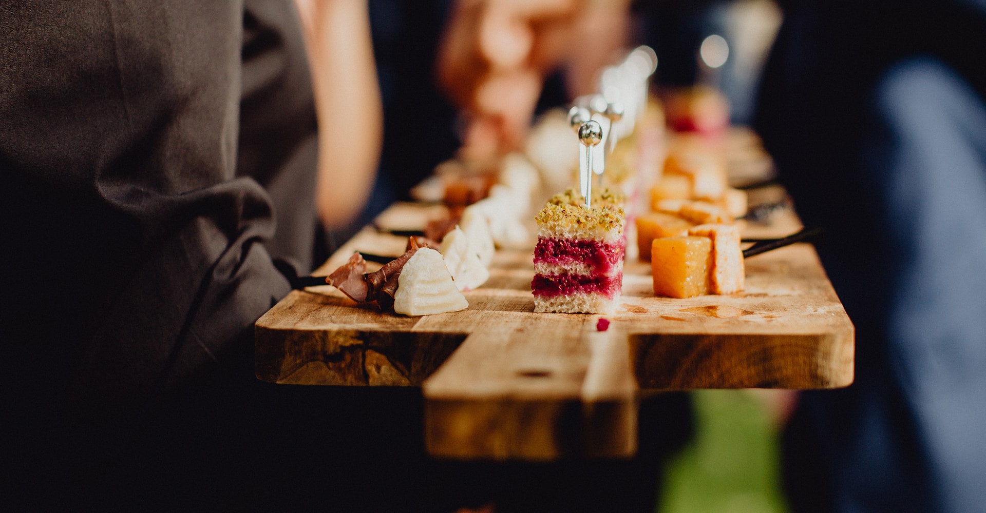 Plateau traiteur en bois. Service d'amuse-bouches : canapés betterave-pistache, cubes de fromage, charcuterie et bouchées blanches.