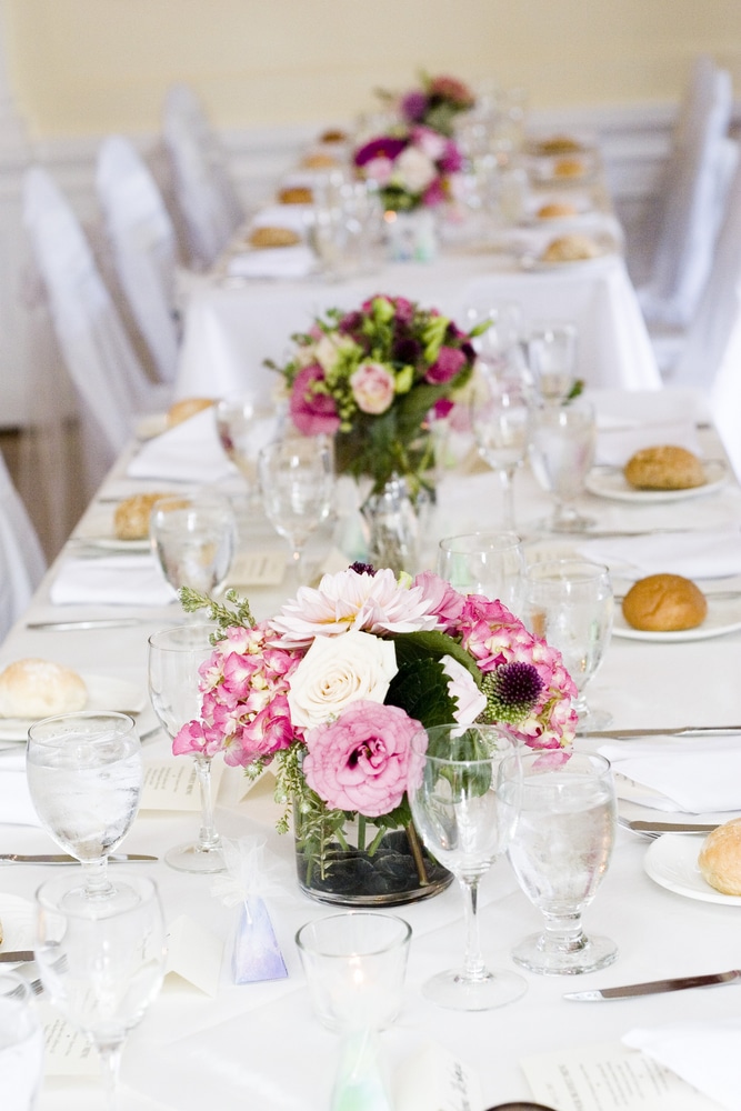 Longue table de réception élégante dressée. Centres de table bas de roses, hortensias et dahlias roses vifs. Nappes blanches, verrerie, pains individuels.